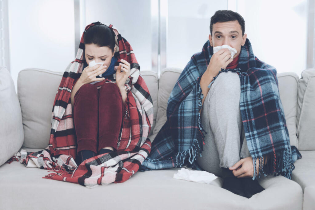 The couple is sitting on the couch wrapped in blankets. Man and woman are sick. The man flies, the woman also blows her nose into a napkin