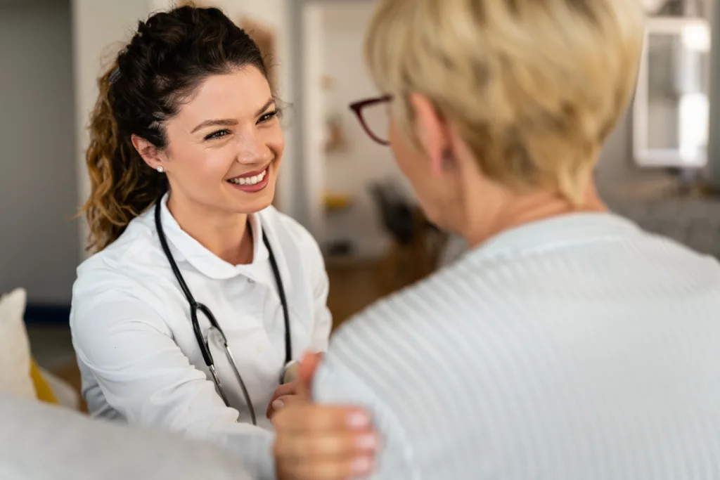 Happy smiling female doctor at home visit talking to unrecognizable senior blonde woman. Support, trust and empathy during counseling or therapy with nurse at home.
