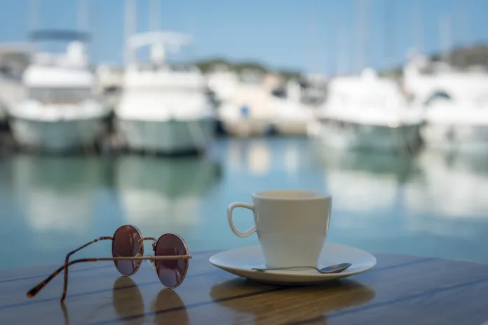 White cup with saucer, sunglasses on a table on the background of blurred boat harbor. Summer vacation concept.