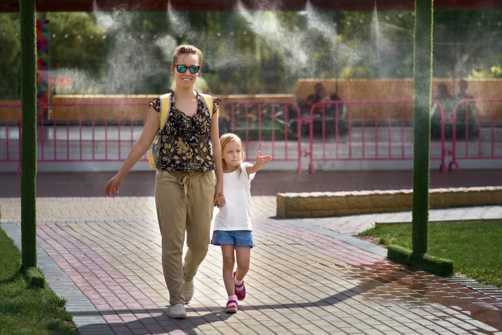 Young mother with her daughter in hot weather, cool off at the gates of climate control in the water park. Aerosol spraying of water jets, spraying of cooling water. Air conditioning nozzle.