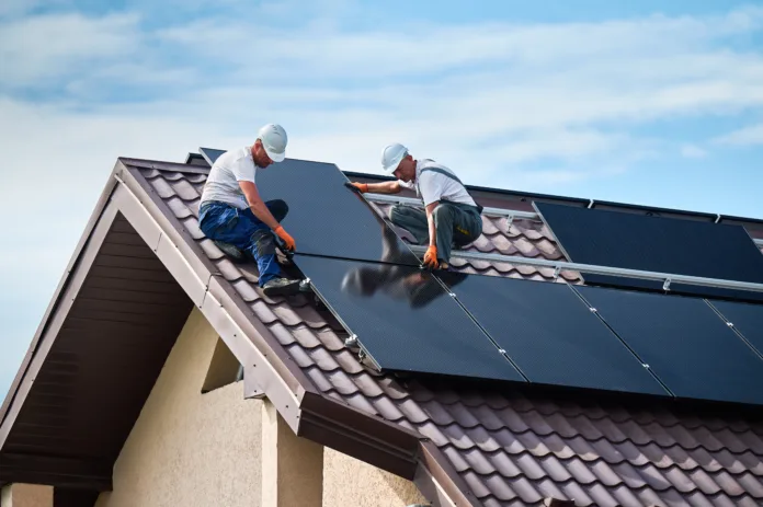 Workers building solar panel system on rooftop of house. Two men installers in helmets installing photovoltaic solar module outdoors. Alternative, green and renewable energy generation concept.