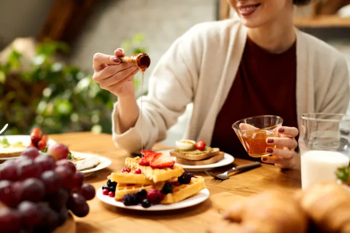 Close-up of woman pouring honey on waffles while eating breakfast at dining table.