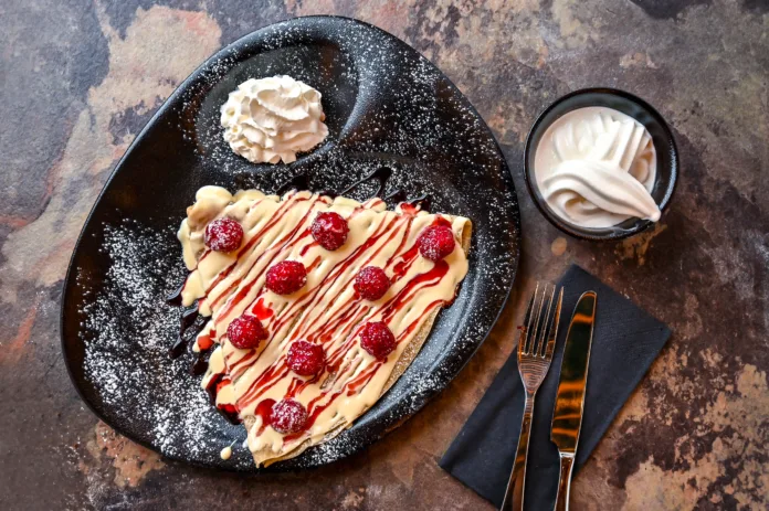 White Chocolate And Raspberry Crepe with whipped cream, knife and fork served in dish isolated on dark background closeup top view of cafe baked dessert food