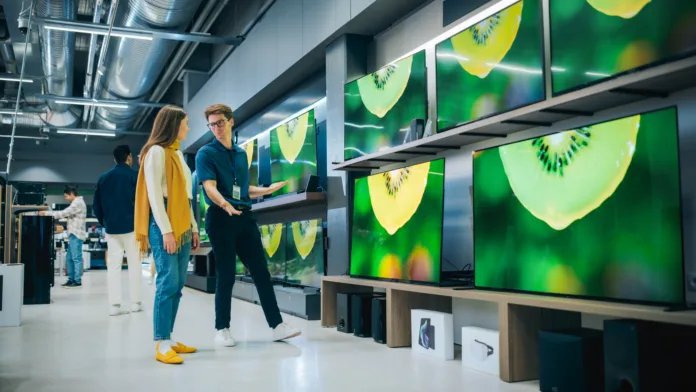 Young Female Shopper Having Conversation with Electronics Store Sales Rep. Stylish Lady Explores Sleek Flat-Screen TV Options. Customer Seeking Impressive Television Solutions in Modern Retail Store