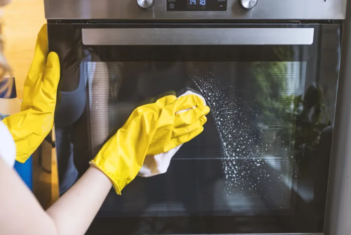 Close up of female hand with yellow protective gloves cleaning oven door. young smiling woman in protective glove with rag cleaning oven. Girl polishing kitchen. People, housework, housekeeping, cleaning concept