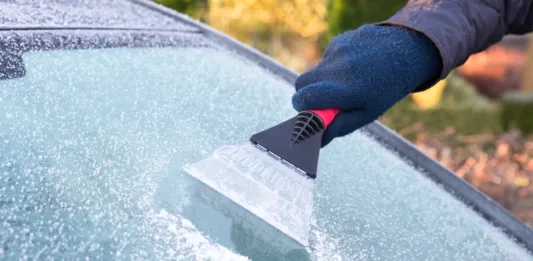 Hand wearing blue glove scratching ice from car window. The ice scraper is an extension of the arm. Part of the windshield is ice free. Only part of the window is visible. Symbol or concept of winter season, cold, frost, weather, sight.